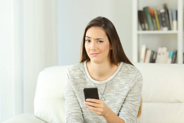 Suspicious woman holding phone looking at you Front view portrait of a suspicious woman holding phone looking at you sitting on a sofa in the living room of a house interior suspicion stock pictures, royalty-free photos & images