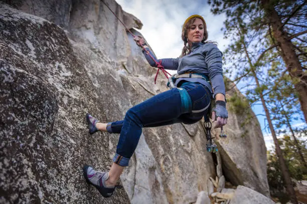 A mid adult caucasian woman rappelling down a rock face, slightly higher than the camera. She is looking down at the ground.
