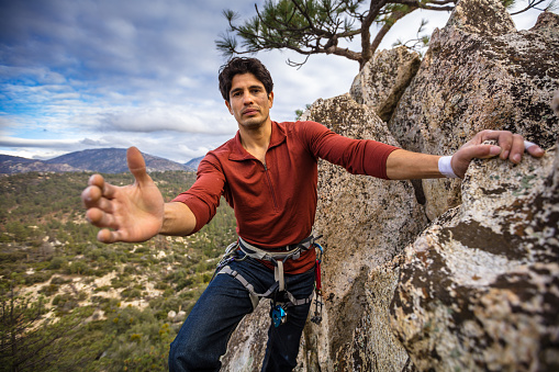 A Latino man in his 30s, wearing a harness full of carabiner clips, holds a hand out to the camera.