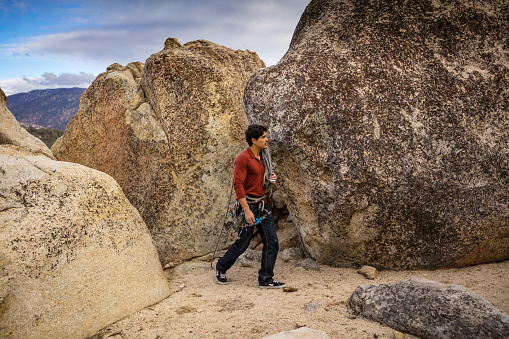A Latino man in his 30s, wearing a harness full of carabiner clips and with a rope looped over his shoulders, walking between large boulders.