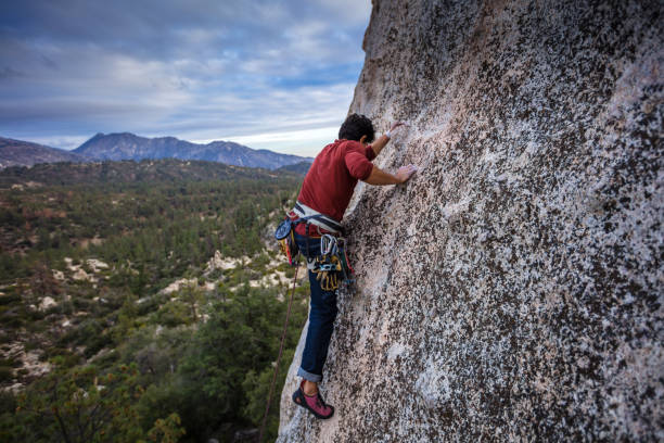 A Latino man in his 30s, wearing a harness full of carabiner clips is lead climbing up a granite rock face in the Angeles National Forest.