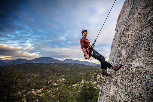 A Latino man in his 30s, wearing a harness full of carabiner clips is abseiling a granite rock face in the Angeles National Forest.