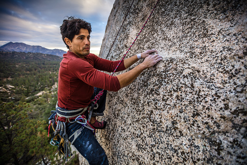 A Latino man in his 30s, wearing a harness full of carabiner clips, his hands covered in chalk, inches along a rock face in the Angeles National Forest, California.