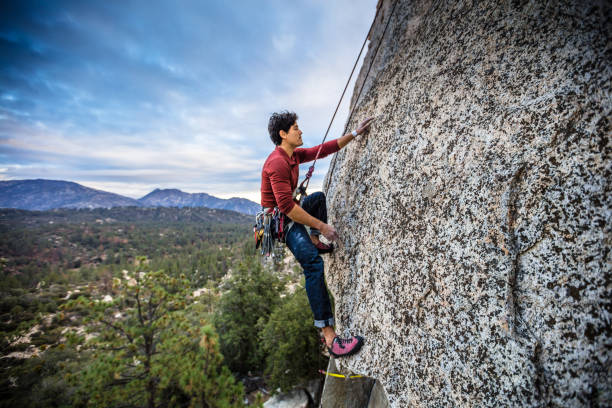 A Latino man in his 30s, wearing a harness full of carabiner clips, his hands covered in chalk, climbing up a rock face in the Angeles National Forest, California.