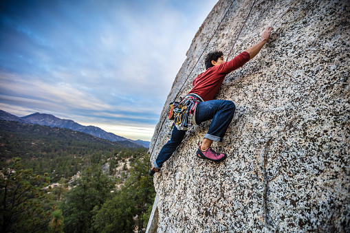 A Latino man in his 30s, wearing a harness full of carabiner clips, his hands covered in chalk, climbing up a rock face in the Angeles National Forest, California.