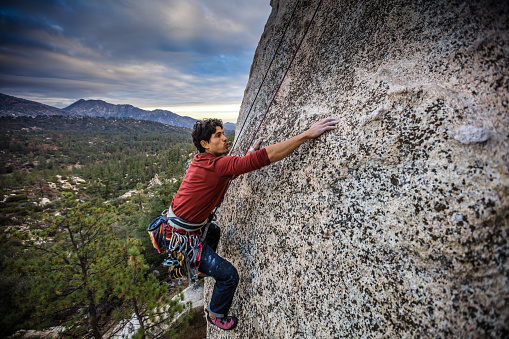 A Latino man in his 30s, wearing a harness full of carabiner clips, his hands covered in chalk, inches along a rock face in the Angeles National Forest, California.