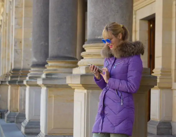 Blonde girl with sunglasses, standing beside pillars, texting.