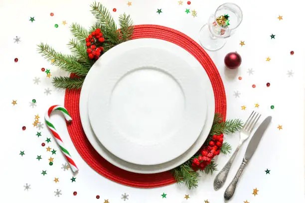 Photo of Christmas table setting with white dishware, silverware and red decorations on white background. Top view.