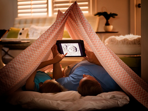 Father and daughter under home made tent made out of bed sheet using digital tablet.