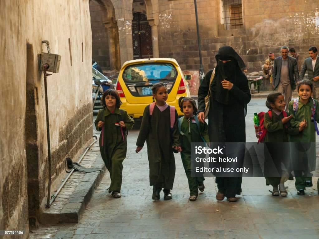 Woman in black dress with small children A woman wearing a black Arabic traditional dress, burka, is walking along a narrow street with a group of young children, schoolchildren. Sanaa is capital city of Yemen. Yemen Stock Photo