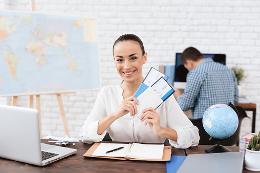 The travel agent keeps tickets for the plane in the travel agency. She offers them to clients. She smiles. On the table she has a toy plane, a laptop.