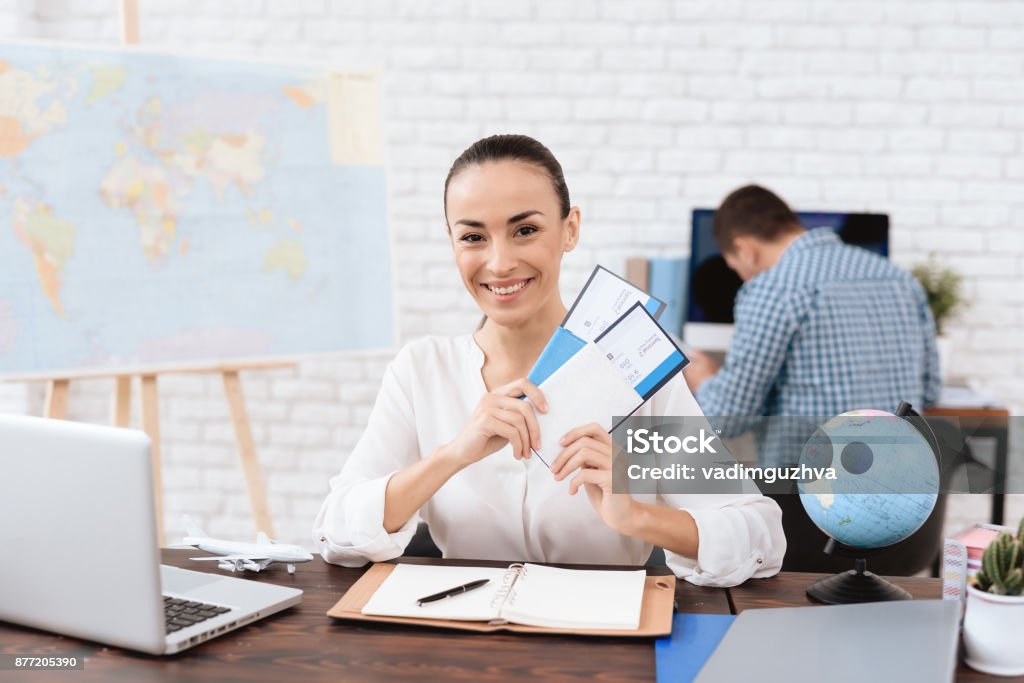 The travel agent keeps tickets for the plane in the travel agency. The travel agent keeps tickets for the plane in the travel agency. She offers them to clients. She smiles. On the table she has a toy plane, a laptop. Travel Agency Stock Photo
