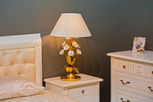 Luxury royal white bedroom in antique style. Bedside table with a chic lamp.