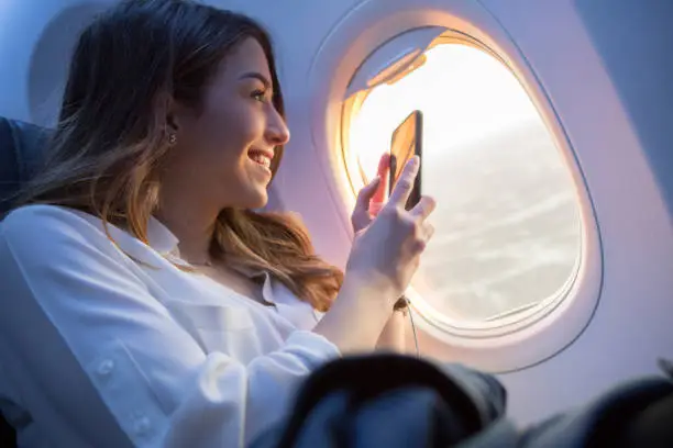 A smiling young woman sits in the window seat of a commercial airliner as it flies through the air.  She is aglow as she looks out the window and takes a picture of a sunset with her smart phone.