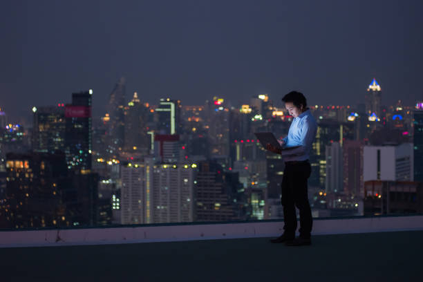 istock Business man standing while working late by computer laptop on rooftop in the city 877257320