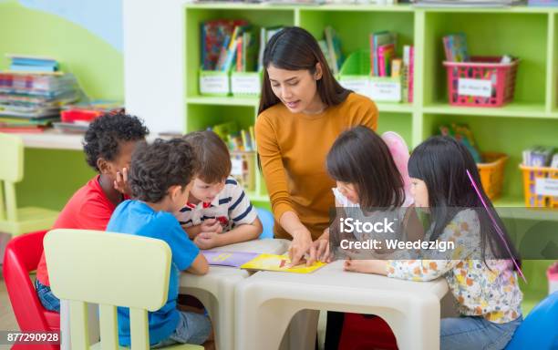 Asian Female Teacher Teaching Mixed Race Kids Reading Book In Classroom Kindergarten Pre School Concept Stock Photo - Download Image Now