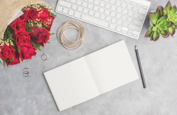 Red roses on grey table with white keyboard, open notebook and boho jewelry stock photo