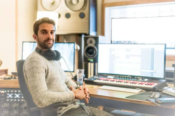 Portrait of confident man sitting on chair in sound recording studio. Record producer is with headphones at workplace. He is wearing casuals.