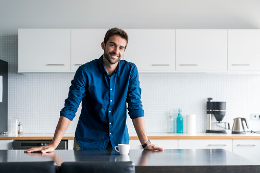 Portrait of smiling mid adult man leaning on kitchen island. Confident handsome male is standing in domestic kitchen. He is wearing casuals at home.
