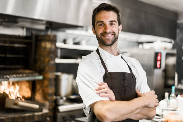 Confident chef standing arms crossed in kitchen Portrait of confident mid adult chef in commercial kitchen. Smiling male cook is standing arms crossed. He is wearing apron. chef stock pictures, royalty-free photos & images