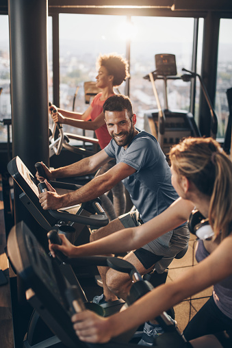 Happy athletic man exercising on exercise bike and talking to his female friend in a health club.