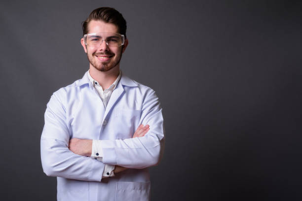 istock Young bearded handsome man doctor wearing protective eyeglasses against gray background 877364524