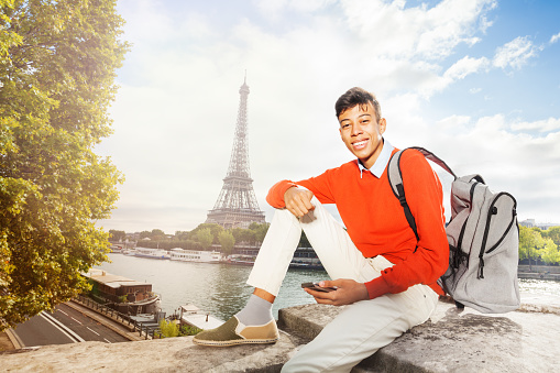 Happy teenage African boy sitting on the embankment of Seine river with the Eiffel Tower on background and using smart phone