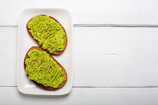 Two rye toast bread with smashed green avocado with lime juice and black pepper on top. On a white plate and wooden background. Horizontal copy space.