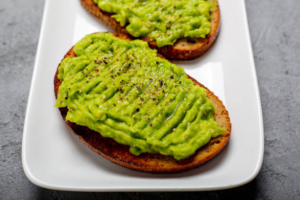 istock Two rye toast bread with smashed green avocado with lime juice and black pepper on top. On a white plate and grey concrete background. Macro close up 877489826