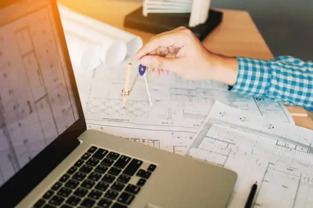 Photo of Architect working on blueprint in workplace with laptop and drawing compass.