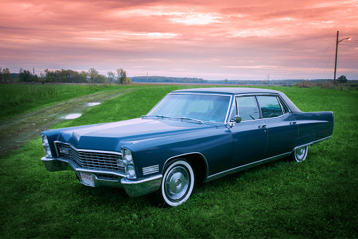 Parked in a field at sunset, with green grass and a purple pink sky wrapping the vehicle in an evening glow.  An old car with its manufacturer’s blue paint, pimped up to drive and look like new.  Three quarter view of the car with its front grill.