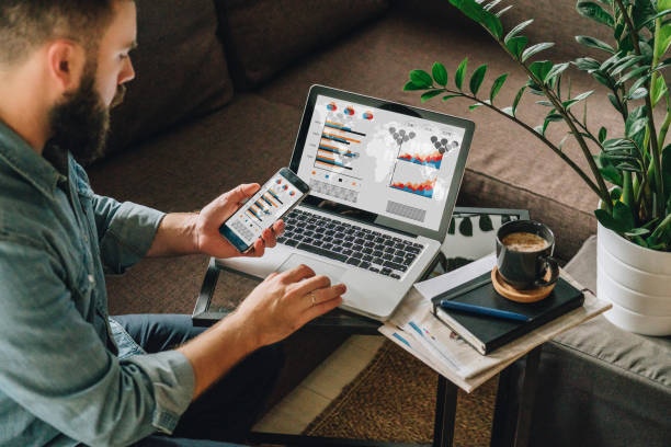 Man, entrepreneur, freelancer sits at home on couch at coffee table, uses smartphone, working on laptop with graphs