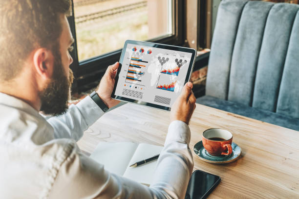 istock Back view. Young bearded businessman in white shirt is sitting at table, using tablet computer with graphs and charts 877748492