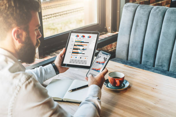 istock Back view.Young bearded businessman in white shirt is sitting at table,using smartphone,tablet computer with graphs 877748596