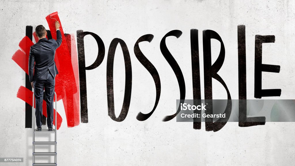 A businessman stands on a stepladder and hides the word Impossible written on the wall using a red paint roller. A businessman stands on a stepladder and hides the word Impossible written on the wall using a red paint roller. Business and success. Goals and planning. Impossible made possible. Conquering Adversity Stock Photo