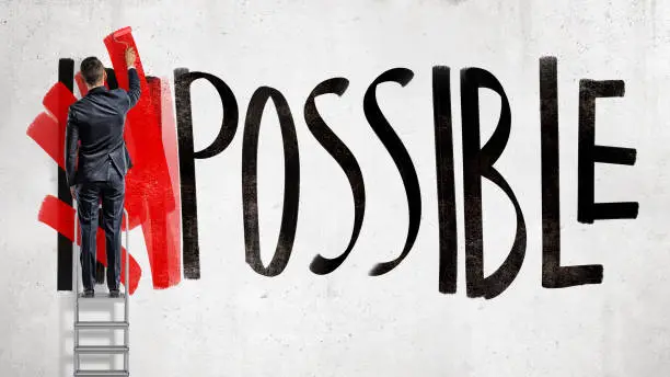 A businessman stands on a stepladder and hides the word Impossible written on the wall using a red paint roller. Business and success. Goals and planning. Impossible made possible.