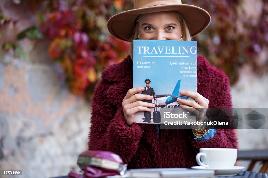 Excited young woman reading journal about journeys I am fold of traveling. Cheerful blond girl is holding magazine near her face and looking at camera with excitement. She is sitting at table in cafe outside Magazine - Publication Stock Photo