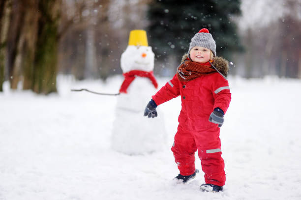 istock Little boy in red winter clothes having fun with snowman 877830016
