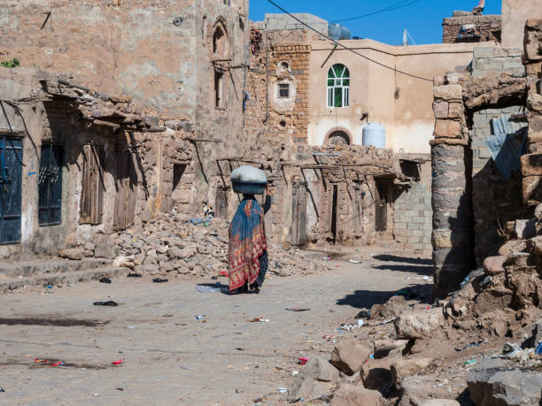 Ruined town in Yemen Outskirts of old part of capital city of Yemen, Sanaa. Buildings ruined and deserted, one women  in traditional clothing walking on dirt road with a basket on her head. Rear view. arabian peninsula photos stock pictures, royalty-free photos & images
