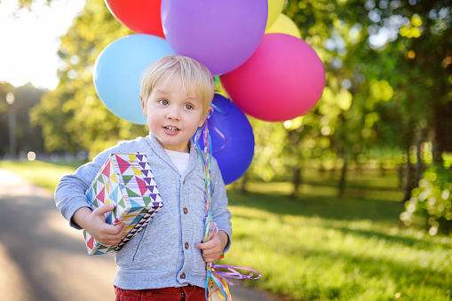 Child going to congratulate a friend on his birthday. Toddler holding bundle of colorful balloons and gift in a festive box. Cute little boy celebrate party with colorful balloons.