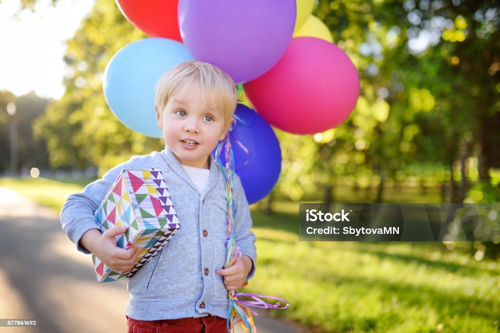 Cute boy holding bundle of colorful balloons and gift in a festive box. Happy Birthday! Child going to congratulate a friend on his birthday. Toddler holding bundle of colorful balloons and gift in a festive box. Cute little boy celebrate party with colorful balloons. Child Stock Photo