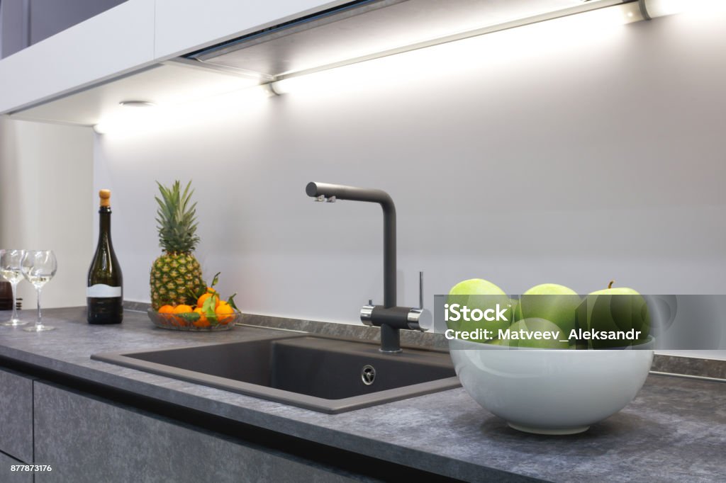 The interior of the modern kitchen is illuminated with a gray stone countertop with a luxury washbasin and mixer, fruit pineapple and tangerines, a bottle with red wine and two glasses. Sink Stock Photo