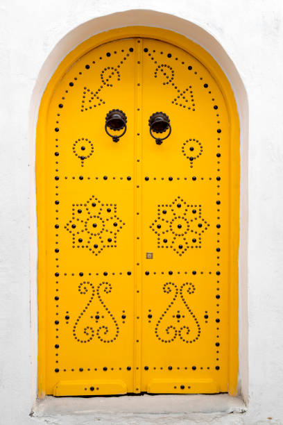 A Caucasian boy is sitting on a step in front of a large, vibrant blue door in Tunis. The setting features traditional Tunisian architecture with intricate patterns and stonework, evoking a sense of exploration and wonder.