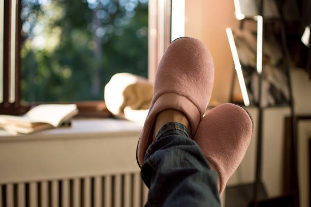 Woman wearing pink slippers with legs up relaxing Relaxing at home - woman wearing jeans and pink slippers taking a break and looking at beautiful day through sunny window at home feet up stock pictures, royalty-free photos & images