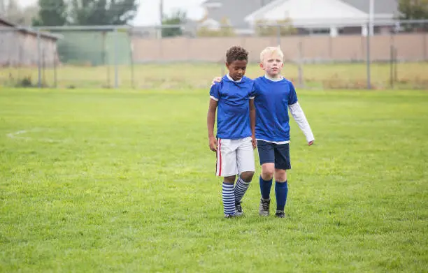 Soccer teammates consoling each other after a tough loss on a soccer field. One boy putting his arm around his friend as they walk off the grassy field. Concept photo of encouragement from friends after disappointment from a loss