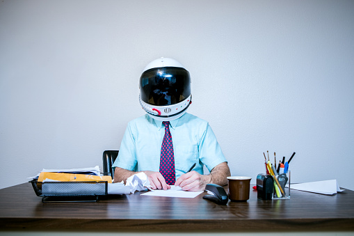 A business man working in the office imagines he is an astronaut, wearing a toy space suit helmet while sitting at his desk.