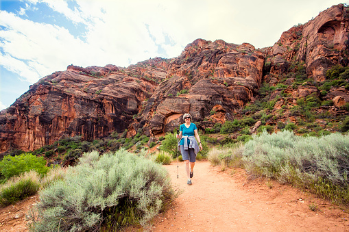 A Happy senior woman hiking in a red rock sandstone canyon. The active, retired woman is enjoying a walk along a scenic trail with vibrant red rock cliffs in the background.