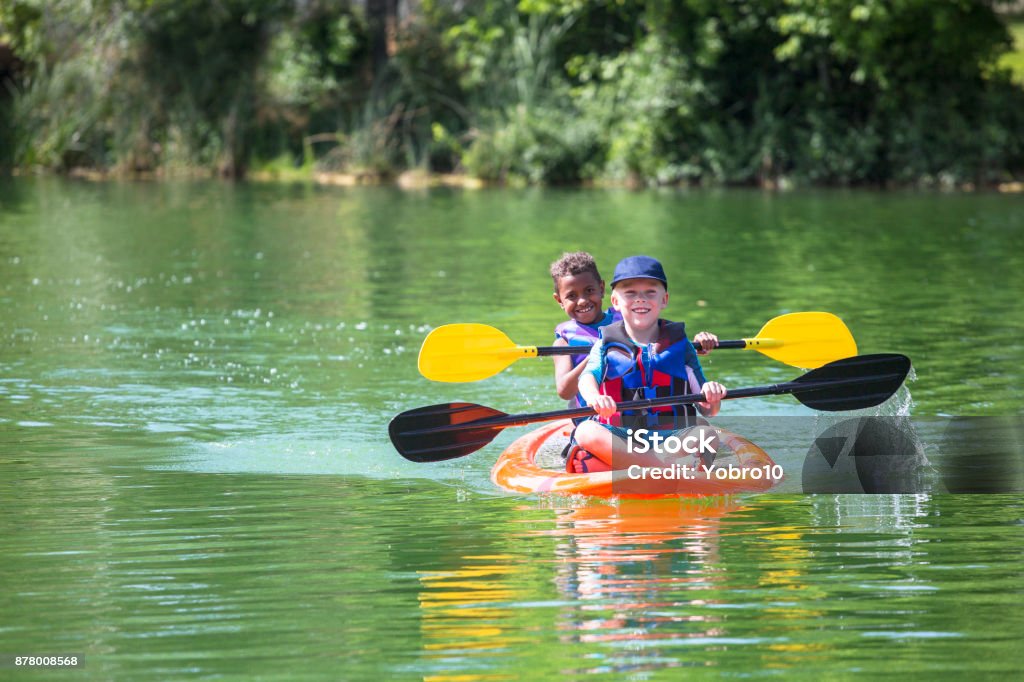 Two diverse little boys kayaking down a beautiful river Two cute diverse young boys kayaking down a beautiful river. Smiling and having fun together on a warm day at summer camp Child Stock Photo