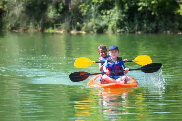 Two cute diverse young boys kayaking down a beautiful river. Smiling and having fun together on a warm day at summer camp