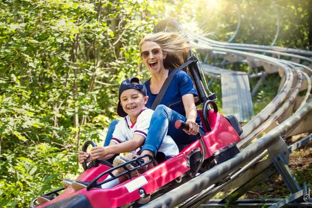 Smiling women and her boy riding downhill together on an outdoor roller coaster on a warm summer day. She has a fun expression as they enjoy a thrilling ride on a red amusement park ride